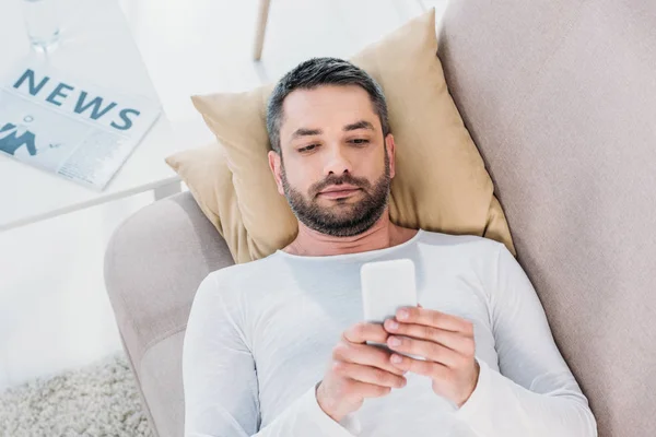 Bel homme allongé sur le canapé et utilisant un smartphone à la maison — Photo de stock