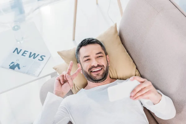 Bel homme souriant couché sur le canapé et prenant selfie sur smartphone à la maison — Photo de stock