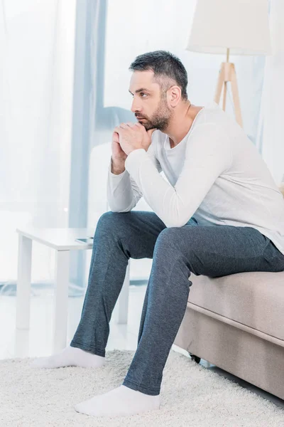 Hombre barbudo guapo sentado en el sofá y apoyando la barbilla con las manos en casa - foto de stock