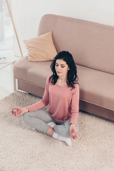Vista en ángulo alto de una hermosa mujer en Lotus Pose practicando meditación en casa en la sala de estar - foto de stock