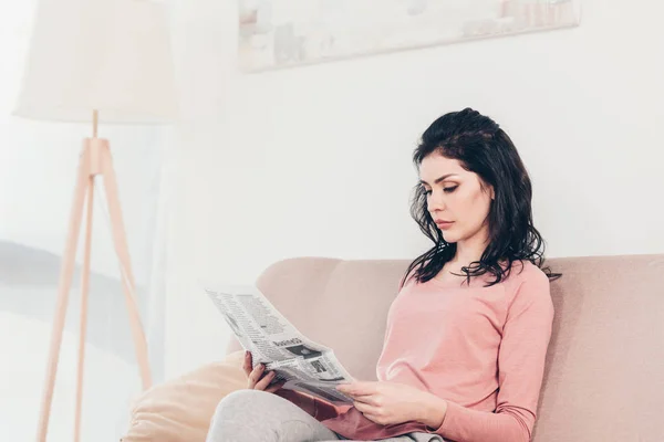 Beautiful serious woman sitting on couch and reading newspaper at home — Stock Photo