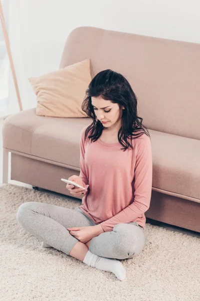 Красива жінка сидить на килимі і використовує смартфон у вітальні вдома — стокове фото