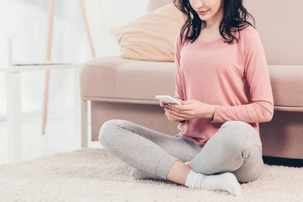 Обрізаний вид жінки, що сидить на килимі і використовує смартфон вдома — стокове фото