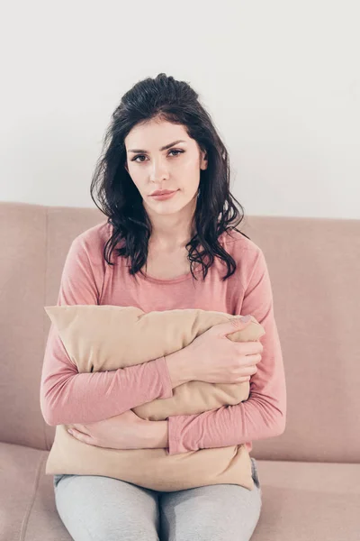 Femme déprimée assise sur le canapé, regardant la caméra et étreignant l'oreiller à la maison — Photo de stock