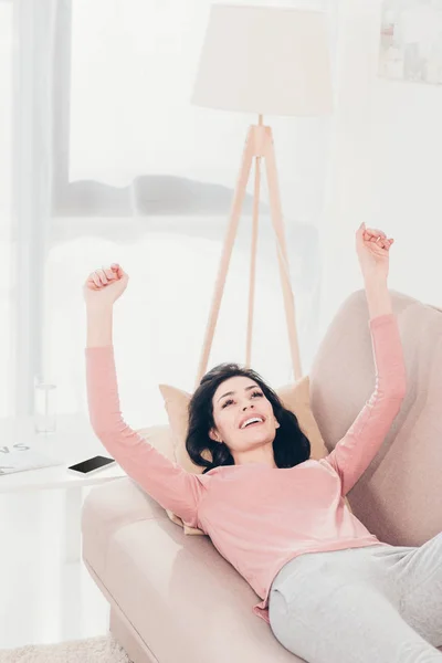 Красивая улыбающаяся женщина с поднятыми руками, отдыхающая дома на диване — стоковое фото
