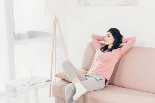 Hermosa mujer con las manos detrás de la cabeza sentado en el sofá en casa con espacio de copia - foto de stock