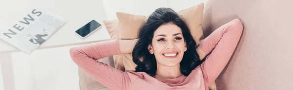 Plano panorámico de hermosa mujer sonriente relajándose en el sofá cerca de la mesa con el periódico y el teléfono inteligente en casa - foto de stock