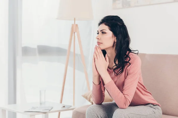 Schöne Frau, die auf dem Sofa sitzt, bitte Geste macht und zu Hause betet — Stockfoto