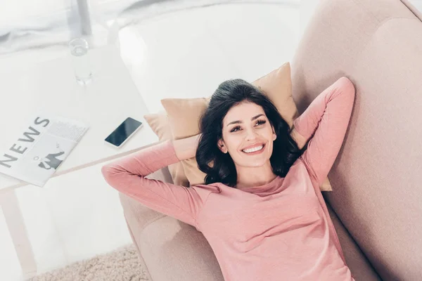 Красива усміхнена жінка розслабляється на дивані біля столу з газетою та смартфоном вдома — стокове фото