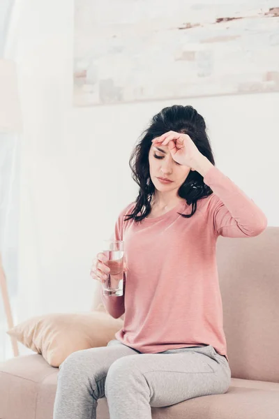 Traurige Frau mit Kopfschmerzen, die auf der Couch sitzt, den Kopf berührt und zu Hause ein Glas Wasser hält — Stockfoto