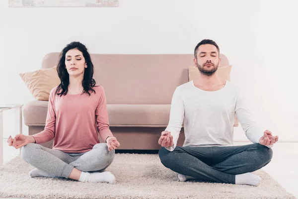 Woman and man with eyes closed sitting in Lotus Pose and meditating at home — Stock Photo