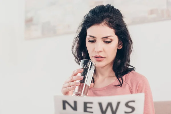 Hermosa mujer sosteniendo un vaso de agua y leyendo el periódico en casa - foto de stock