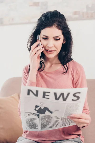 Schöne Frau, die zu Hause auf dem Smartphone spricht und Zeitung liest — Stockfoto