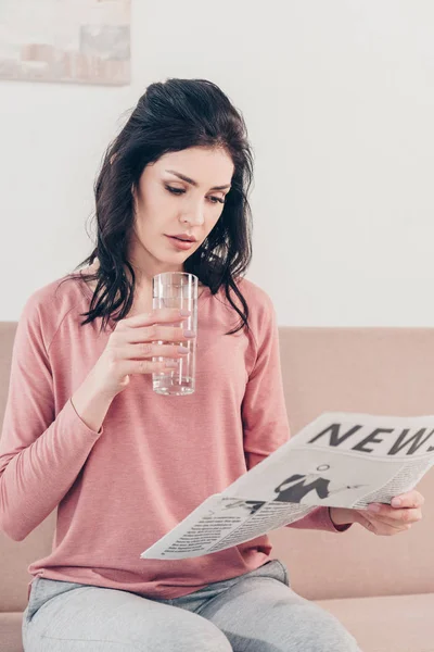 Beautiful woman holding glass of water and reading newspaper at home — Stock Photo