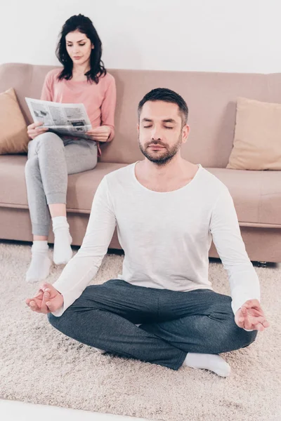 Frau sitzt auf Couch und liest Zeitung, während Mann zu Hause in Lotus-Pose meditiert — Stockfoto