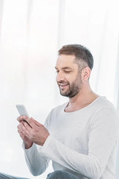 Bell'uomo sorridente in abiti casual utilizzando smartphone a casa — Foto stock