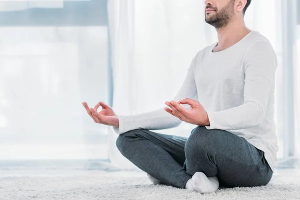 Cropped view of man sitting on carpet in Lotus Pose and meditating at home — Stock Photo