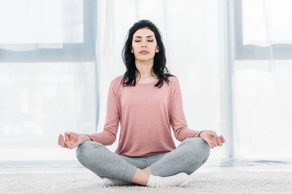 Hermosa mujer con los ojos cerrados en Lotus Pose practicando meditación en la sala de estar en casa - foto de stock