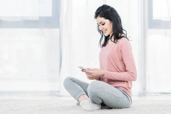 Hermosa mujer sonriente sentada en la alfombra y el uso de teléfono inteligente en casa con espacio para copiar - foto de stock