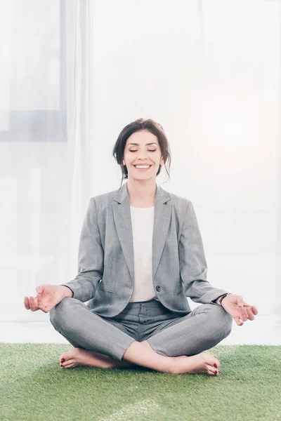 Beautiful smiling businesswoman in suit meditating while sitting on grass mat in Lotus Pose — Stock Photo