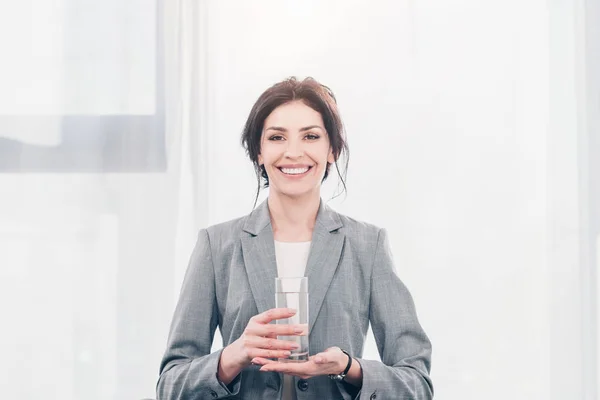 Hermosa mujer de negocios sonriente en traje sosteniendo un vaso de agua y mirando a la cámara - foto de stock