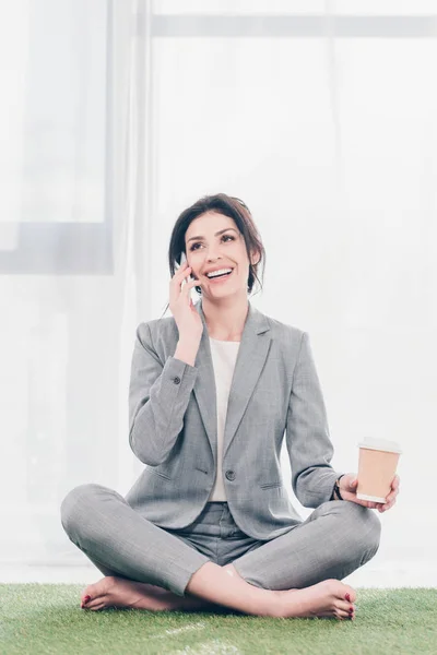 Beautiful smiling businesswoman in suit sitting on grass mat, holding coffee to go and talking on smartphone — Stock Photo