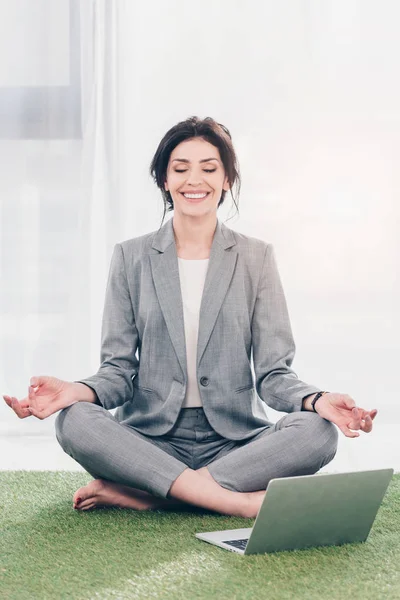 Smiling businesswoman in suit meditating while sitting on grass mat in Lotus Pose near laptop — Stock Photo