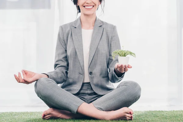 Обрізаний вид посміхаючись ділової жінки в костюмі медитації на траві мат в Lotus позі з вазон — стокове фото