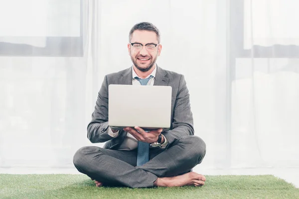 Guapo sonriente hombre de negocios en traje sentado en la alfombra de hierba y el uso de ordenador portátil - foto de stock
