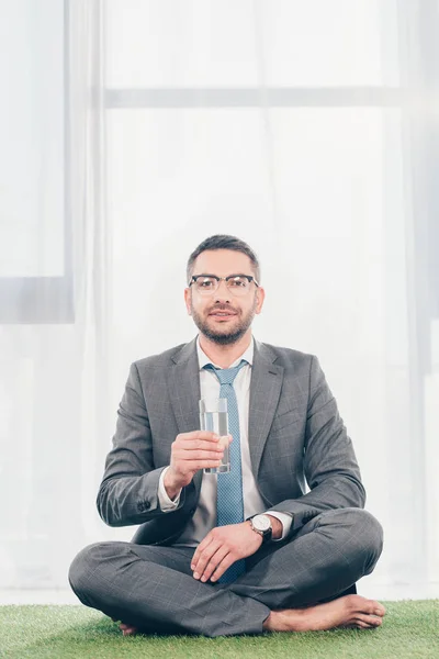 Handsome businessman in suit sitting on grass mat, looking at camera and holding glass of water — Stock Photo