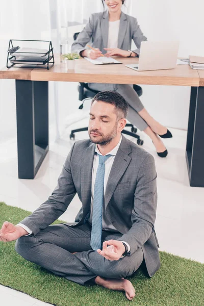 Geschäftsmann meditiert in Lotus-Pose auf Grasmatte, während Geschäftsfrau im Büro am Tisch sitzt — Stockfoto