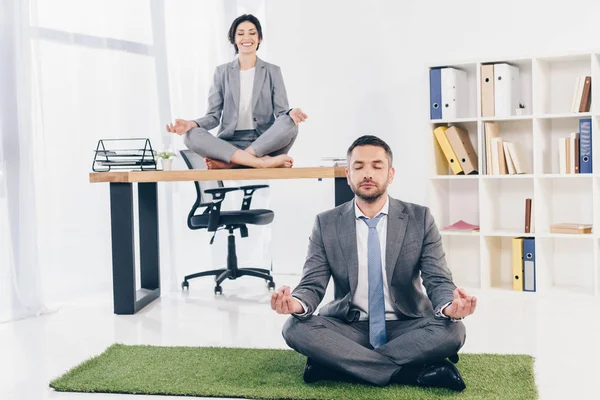 Hombre de negocios meditando en alfombra de hierba mientras que la mujer de negocios sentada en la mesa en Lotus Pose en la oficina - foto de stock