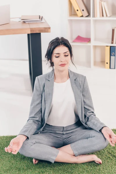 Hermosa mujer de negocios con los ojos cerrados sentado en la alfombra de hierba y meditando en Lotus Pose en la oficina - foto de stock