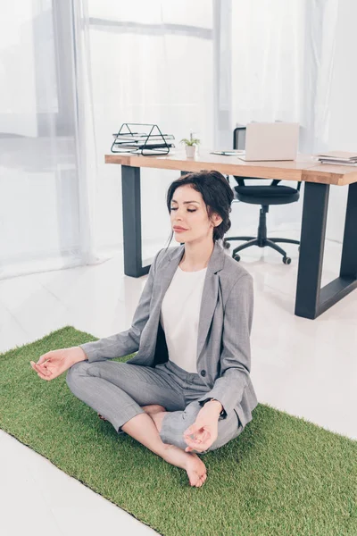 Beautiful businesswoman in suit with eyes closed sitting on grass mat and meditating in Lotus Pose in office — Stock Photo