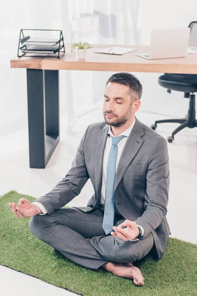 Sentado en la alfombra de hierba en Lotus Pose y meditando en la oficina - foto de stock