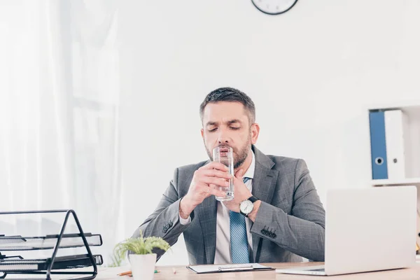 Hombre de negocios guapo en traje sentado en el escritorio, tocando la garganta y el agua potable en la oficina - foto de stock