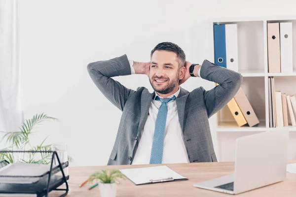 Bell'uomo d'affari sorridente in giacca e cravatta con le mani dietro la schiena seduto alla scrivania in ufficio — Foto stock