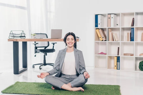 Красивая улыбающаяся деловая женщина в костюме сидит на травяном коврике в Lotus Pose и медитирует в офисе — стоковое фото