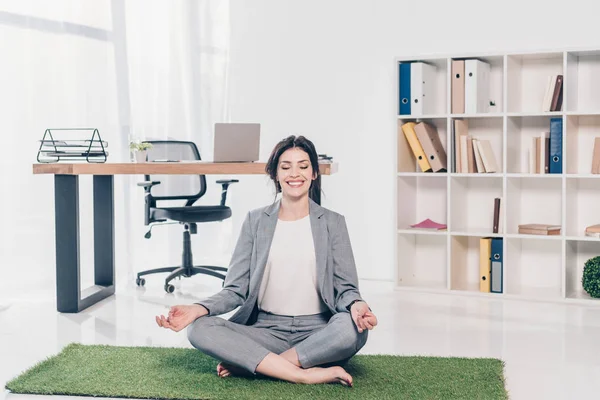 Hermosa mujer de negocios sonriente en traje sentado en la estera de hierba en Lotus Pose y meditando en la oficina - foto de stock