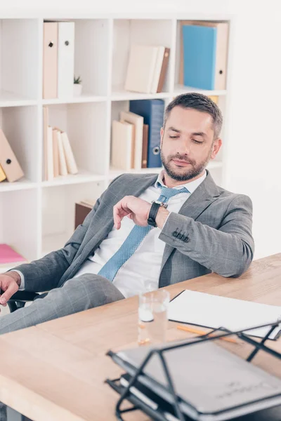 Schöner Geschäftsmann, der am Schreibtisch sitzt, auf die Uhr schaut und im Büro die Uhrzeit überprüft — Stockfoto