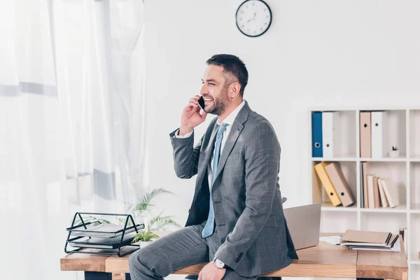 Симпатичный улыбающийся бизнесмен в костюме разговаривает на смартфоне в офисе — стоковое фото