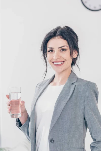Красива усміхнена бізнес-леді в костюмі тримає склянку води і дивиться на камеру — стокове фото