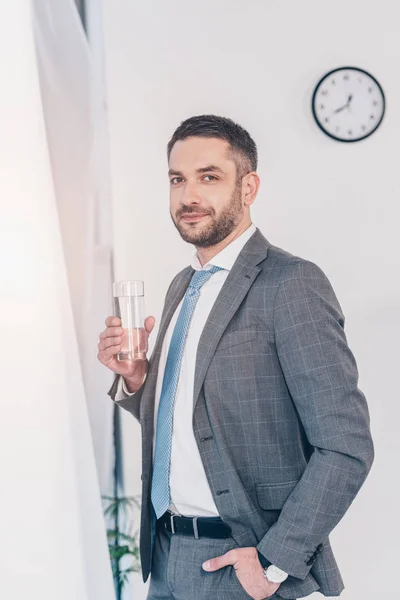 Handsome businessman in suit holding glass of water and looking at camera in office — Stock Photo