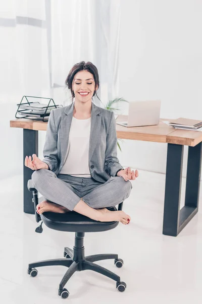 Hermosa mujer de negocios sonriente en traje sentado en la silla y meditando en Lotus Pose en la oficina - foto de stock
