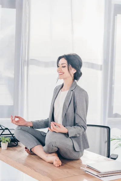 Schöne lächelnde Geschäftsfrau im Anzug meditiert auf dem Tisch in Lotus-Pose im Büro — Stockfoto