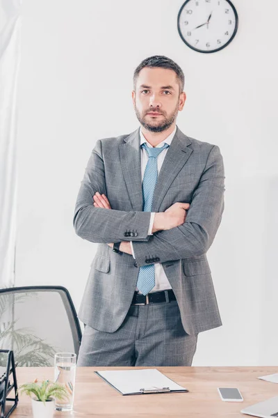 Serious handsome businessman in suit with crossed arms looking at camera in office — Stock Photo