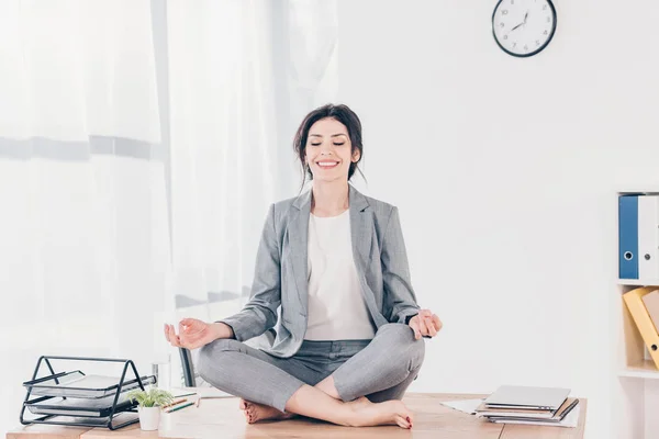 Schöne lächelnde Geschäftsfrau im Anzug sitzt am Schreibtisch und meditiert in Lotus-Pose im Büro — Stockfoto