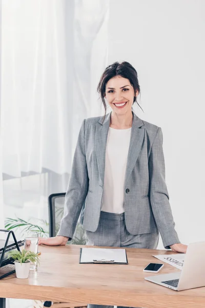 Красивая улыбающаяся деловая женщина в костюме за столом смотрит в камеру в офисе — стоковое фото