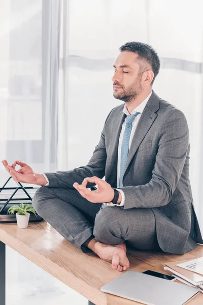 Schöner Geschäftsmann im Anzug meditiert in Lotus-Pose, während er auf dem Bürotisch sitzt — Stockfoto