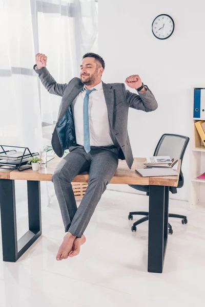 Hombre de negocios guapo en traje con los ojos cerrados sentado en el escritorio y estiramiento en la oficina - foto de stock